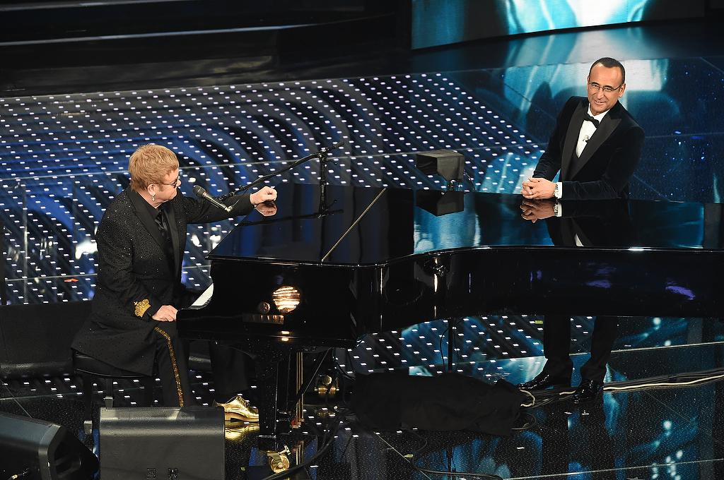 Elton John sul palco di Sanremo 206 con Carlo Conti indossa una scintillante giacca custom made, che riporta sulla schiena le iniziali E.J. in oro, e che il cantante abbina a un paio di stringate della stessa tinta (Fotogramma)