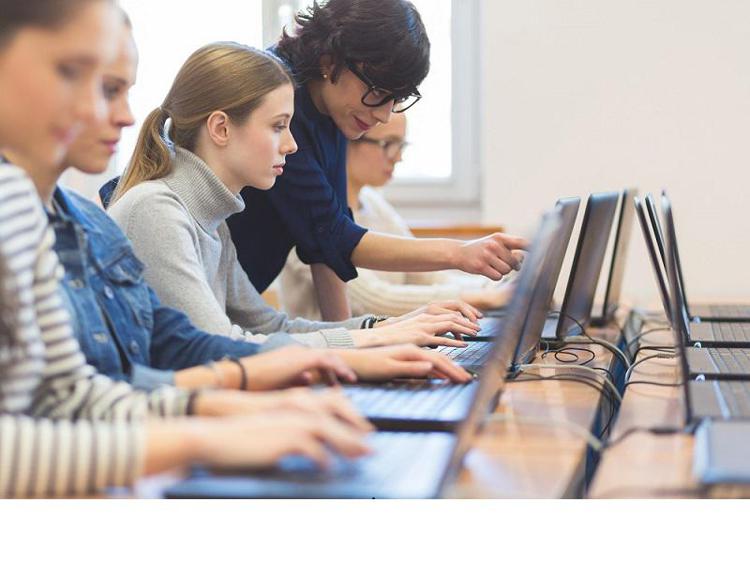 Side view of group of female students coding on laptops in a computer lab. Teacher pointing at the computer screen. - Getty Images